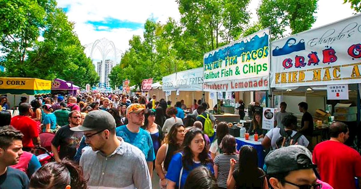 A crowded festival walkway at Bite of Seattle lined with food vendor stalls selling fish tacos, crab, and fried mac and cheese, under trees near Seattle Center's pergola